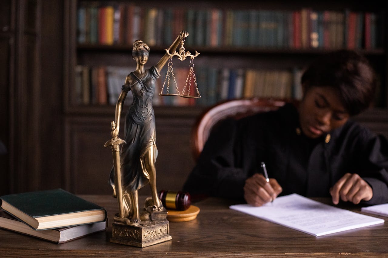 services-01 A focused judge writing on documents beside a Lady Justice statue in an office.